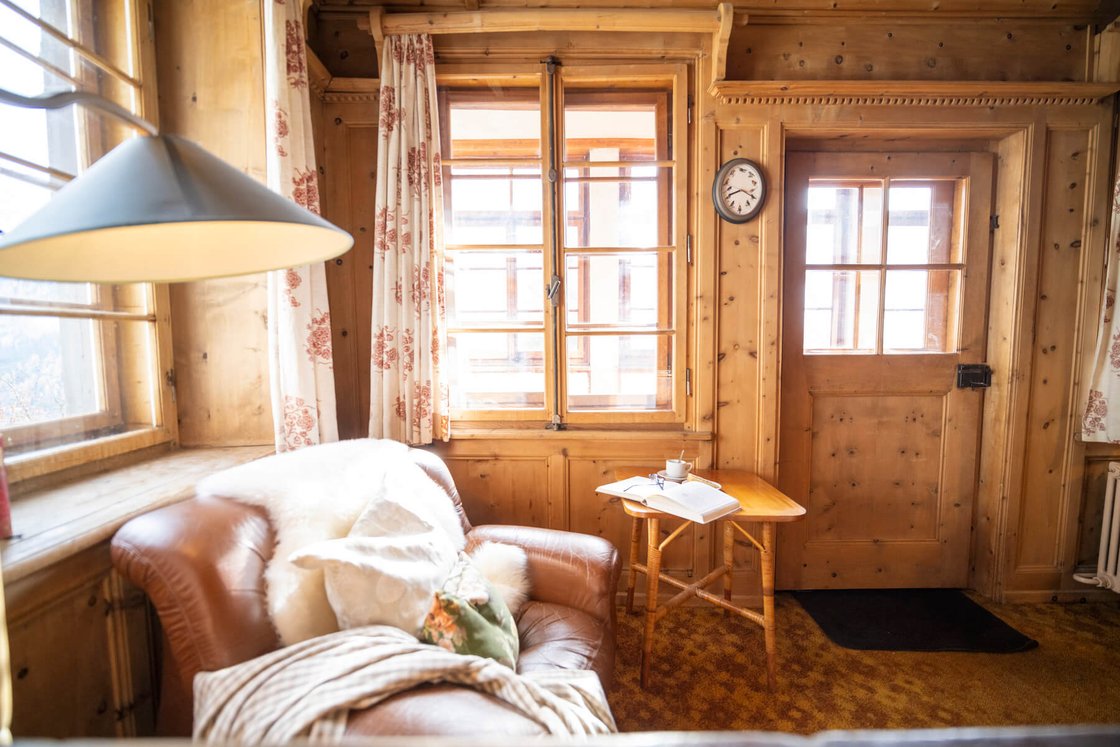 Eine gemütliche Leseecke im La Randulina: ein Ledersessel mit Schaffell, ein Beistelltisch aus Holz mit einem Buch und eine antike Uhr an den alten Holzwänden.