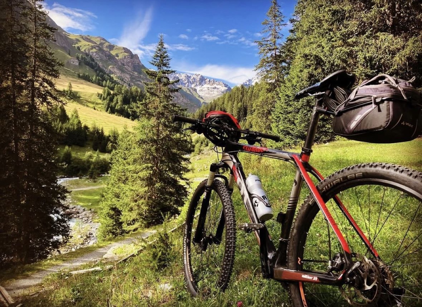 Ein Mountainbike an einem Weg entlang eines Bergbachs, mit verschneiten Gipfeln über dem Tal.