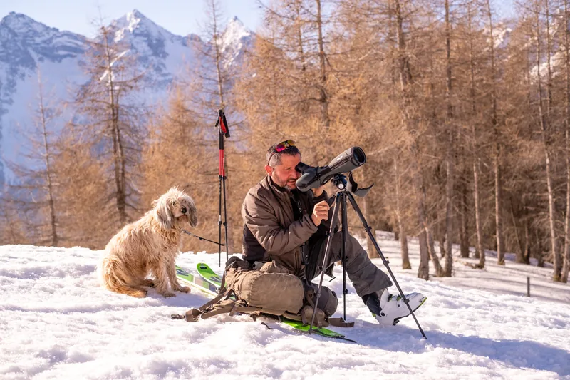 Een man kijkt door een telescoop in de sneeuw, met een hond naast zich en lariksen erachter.