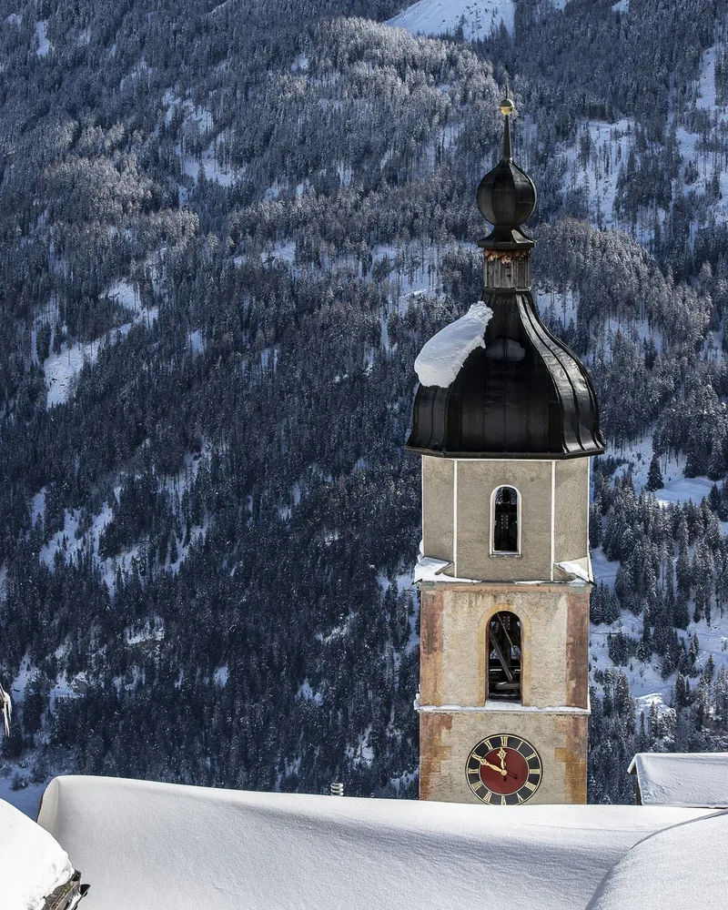 Een kerktoren boven de besneeuwde daken, met beboste bergen erachter.