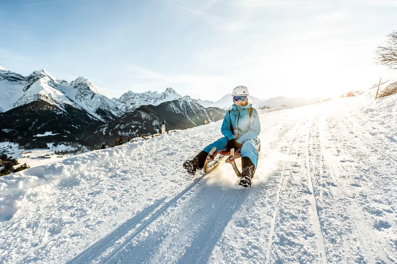 Iemand in een blauw skipak, met witte helm en skibril, op een houten slee op een besneeuwde weg, met besneeuwde bergtoppen en een kerktoren op de achtergrond.