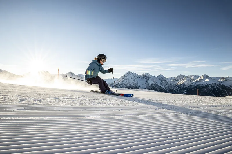 Een skiër maakt een bocht op een vers geprepareerde piste, met de zon laag boven de bergen.